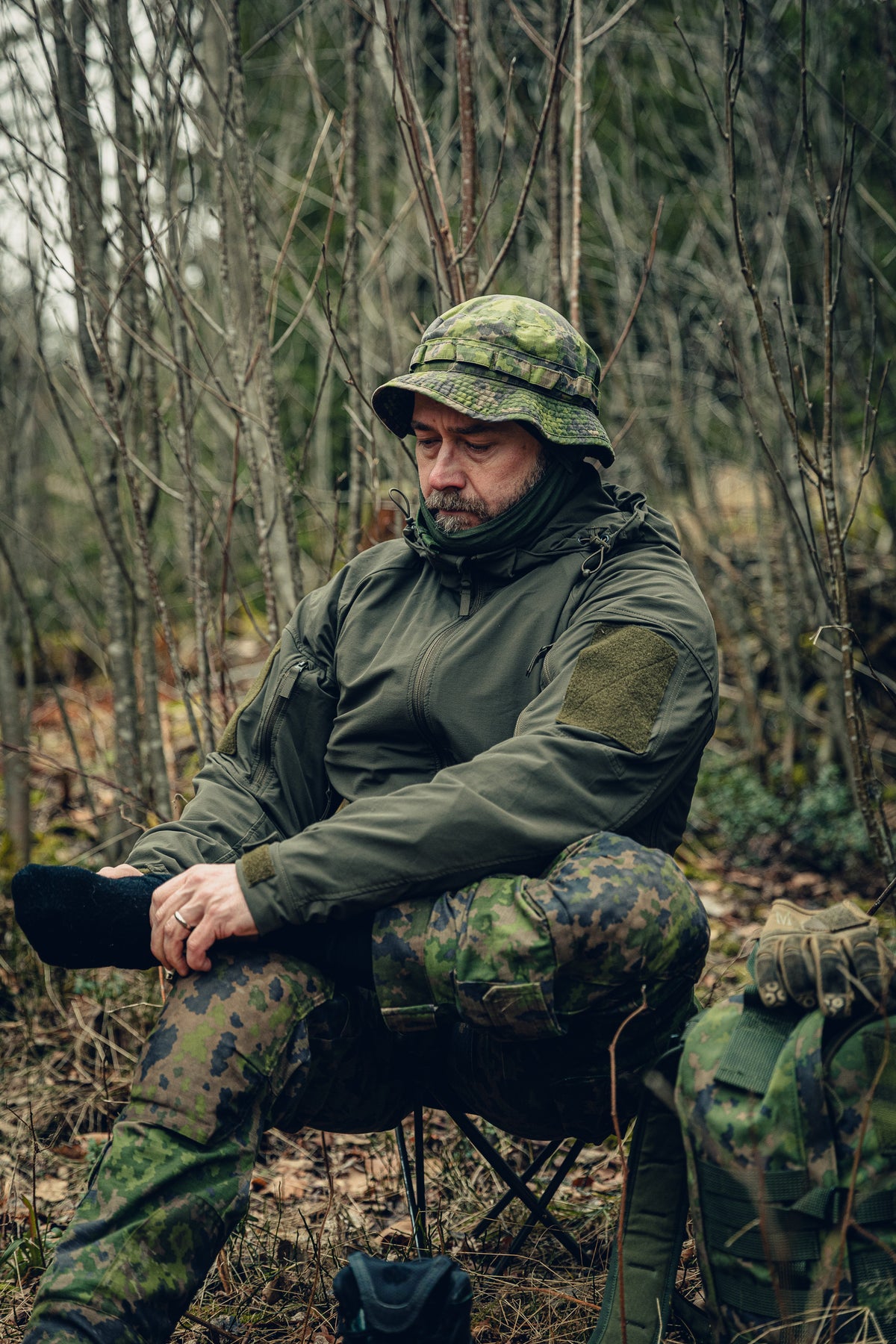 A man sitting on a stool in the forest changing his socks. He is wearing Finnish M05 woodland pants and a boonie hat, a green balaclava pulled down to his chin and a green jacket, and there is a M05 woodland backpack next to him on the ground.