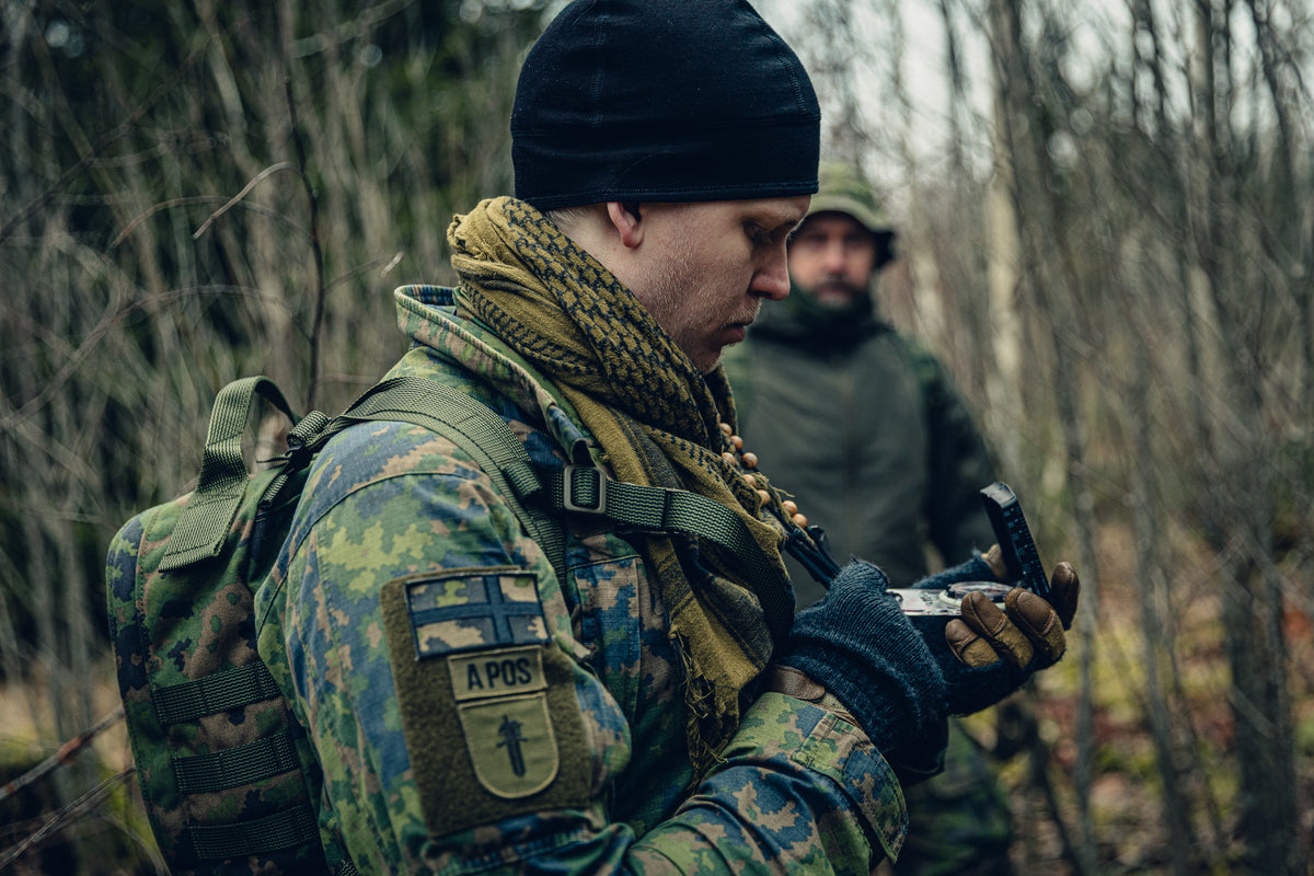 A man looking at a compass in the forest with another man standing in the background. The man in the front is wearing Finnish M05 woodland clothing and a backpack, he has the Finnish flag patch, a blood type patch and a training batch patch on his sleeve, a black beanie and a brown shemagh.