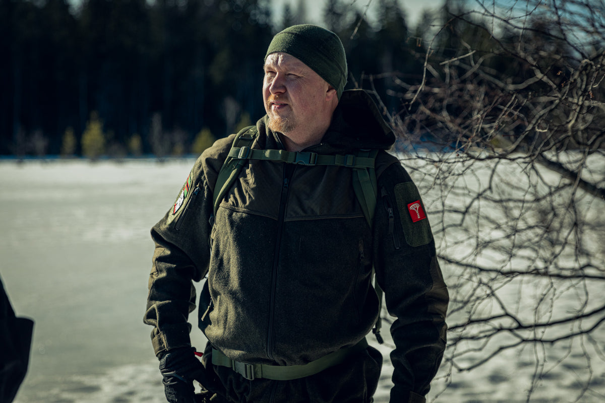 A man squinting his eyes and standing in front of a frozen lake with the sun shining. He is wearing a green jacket, a green beanie and you can see the sternum strap of a backpack.
