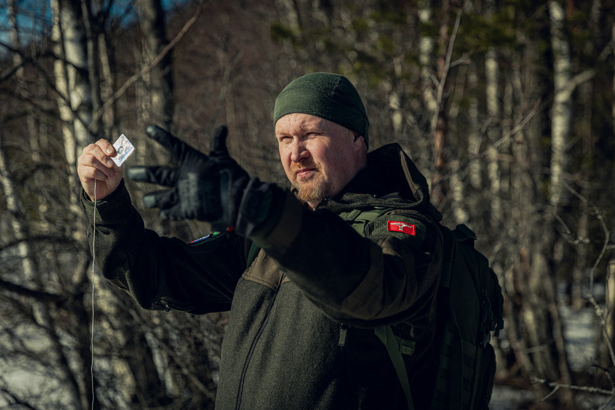 A man wearing a thin green beanie and green wool jacket holding a small mirror in his right hand and spreading out his gloved fingers of his left hand. You can see a slightly snowy but sunny forest in the background.