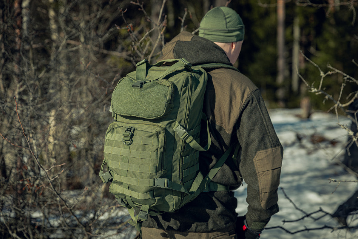A backshot of a man in a snowy forest wearing green clothing and a green backpack. The sun is shining and he seems to be walking away from the camera.