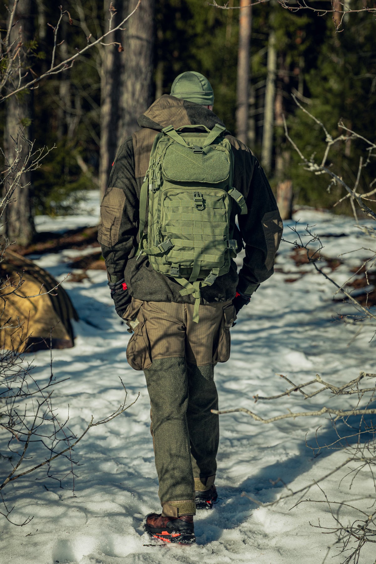 A man in a sunny, snowy forest walking away from the camera. He is wearing a green jacket and pants, a green backpack and a green beanie.