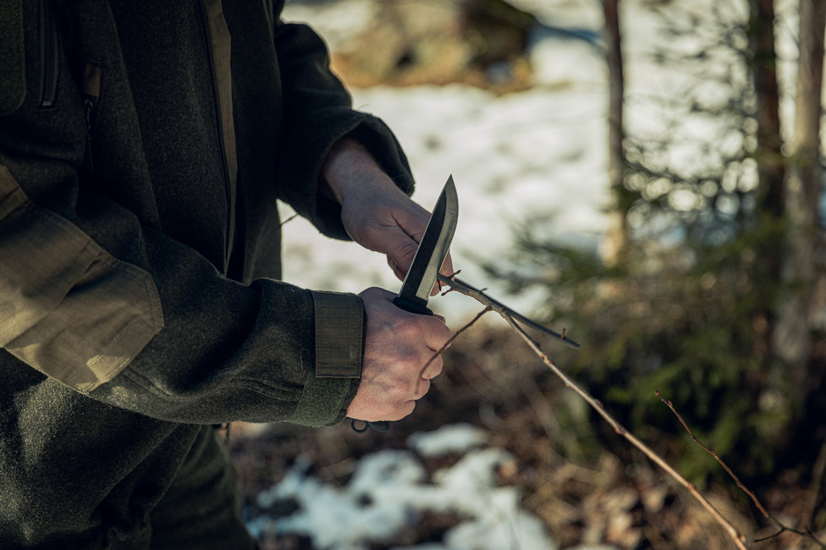 Nahaufnahme von Händen, die ein Messer neben einem dünnen Ast halten. Die Person trägt grüne Wollkleidung, und im Hintergrund sieht man einen leicht verschneiten, aber sonnigen Wald.