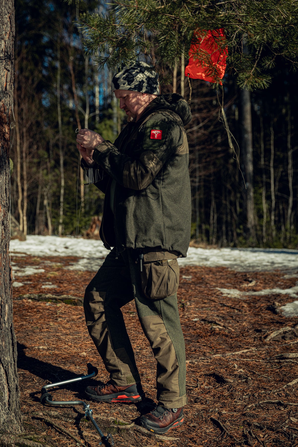 Full-body shot of a man standing next to a pine tree in a sunny forest with some snow on the ground. He is holding a thin thread in his hands, wearing a green jacket and pants and a black-and-white beanie. You can see some equipment on the ground and a bright orange fabric in the tree.