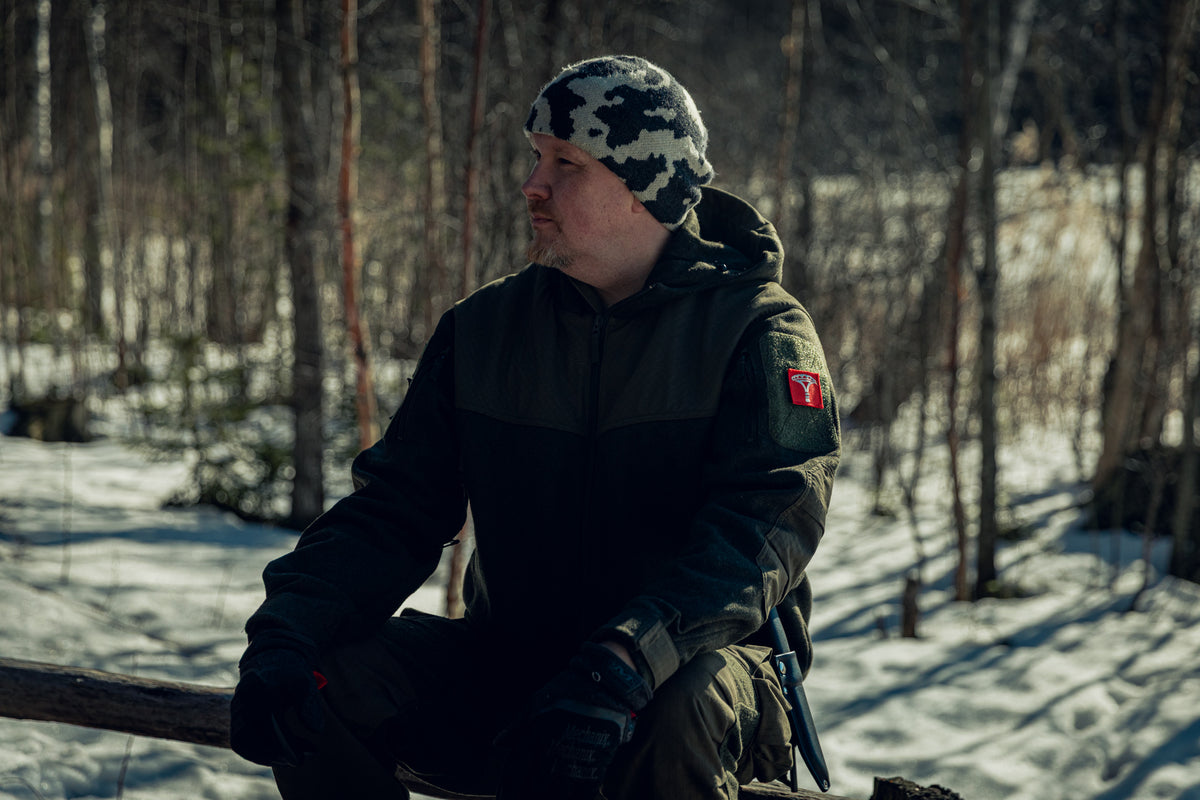 A man sitting in a snowy, sunny forest. He looking to the left and wearing a green woolen jacket with some patches on the sleeve, black gloves, and a snow camo beanie. There is a knife in a black leather sheath on his belt.