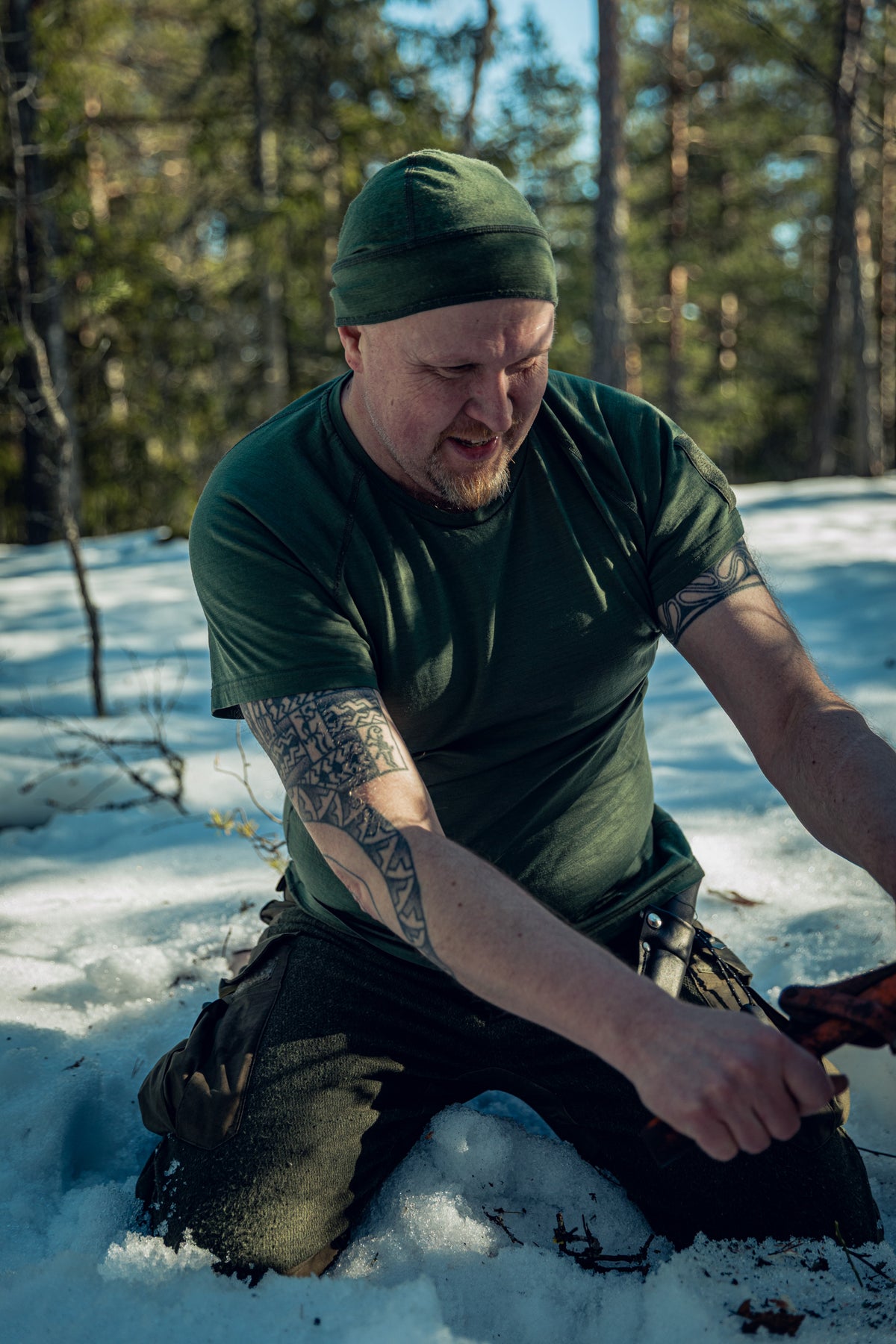 Ein Mann in einem sonnigen, verschneiten Kiefernwald kniet im Schnee und hält eine Axt in den Händen. Er trägt grüne Hosen, ein grünes T-Shirt und eine grüne Mütze. Er hält etwas Ausrüstung in den Händen, vielleicht den Griff einer Säge.
