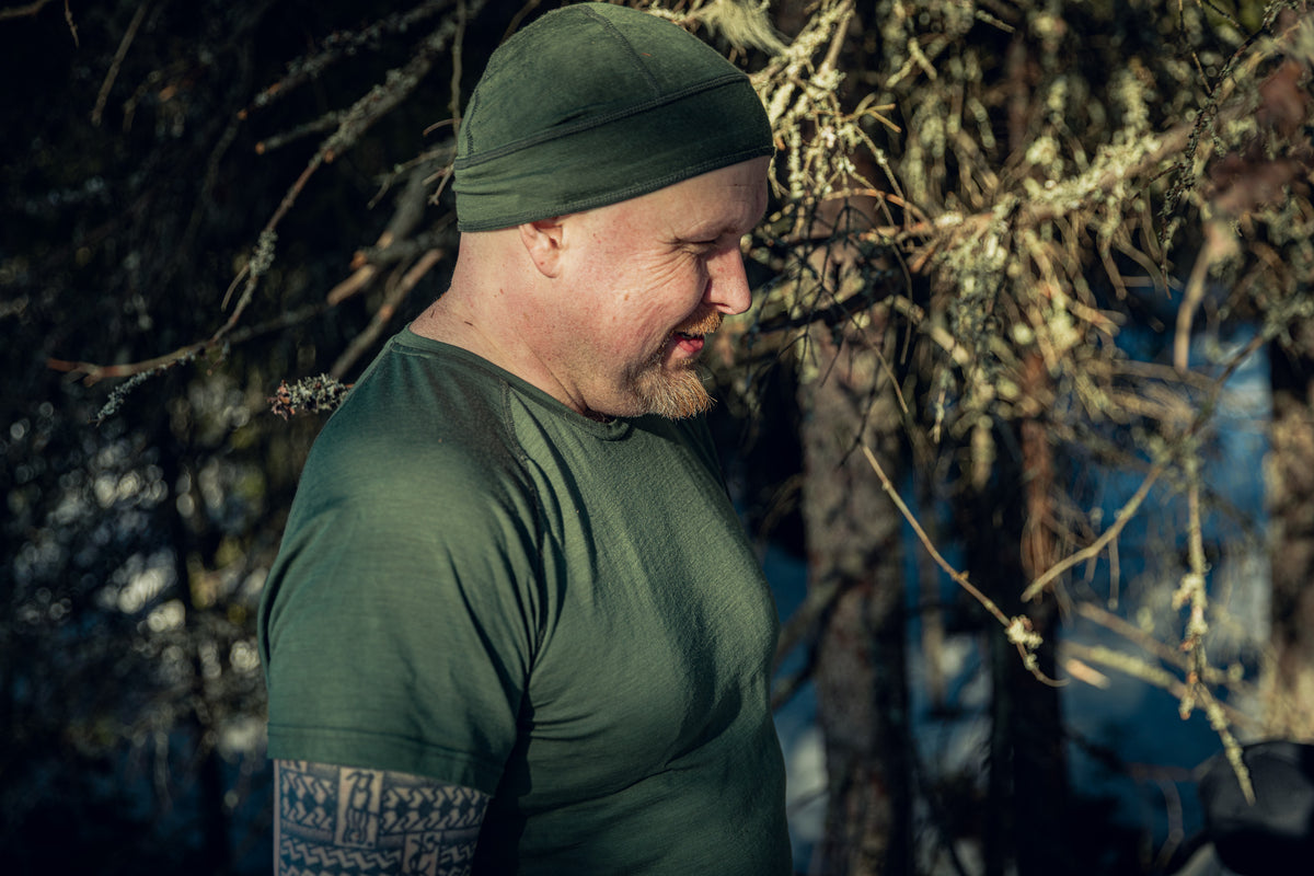 Close-up of a man in a sunny forest with some snow visible on the ground. He is smiling, wearing a green beanie and a green t-shirt.