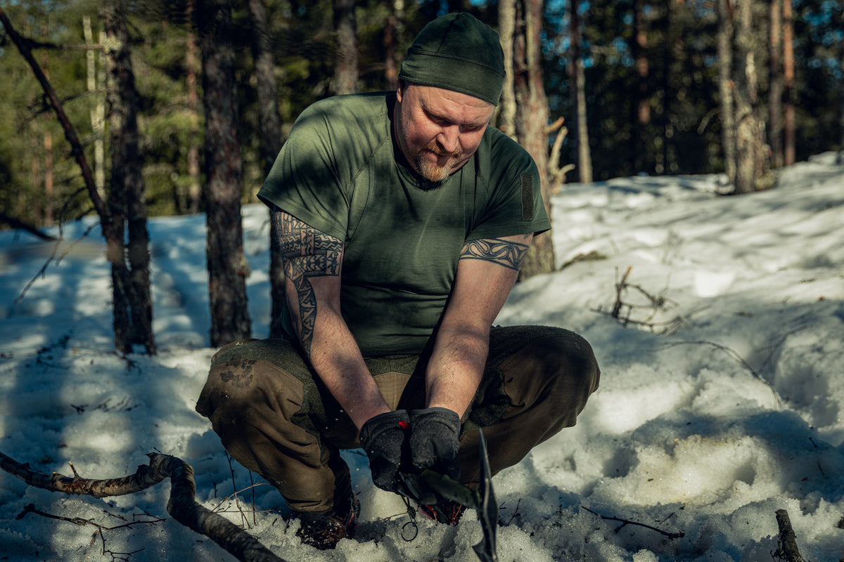 Ein Mann in einem sonnigen, verschneiten Kiefernwald hockt im Schnee und hält eine Axt in den Händen. Er trägt grüne Hosen, ein grünes T-Shirt, schwarze Handschuhe und eine grüne Mütze.