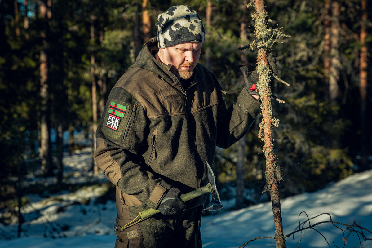 A man in a sunny pine wood forest with snow on the ground, holding a thick branch in one hand and an axe in the other. He is wearing a green woolen jacket with some patches on the sleeve, black gloves and a snow camo beanie.