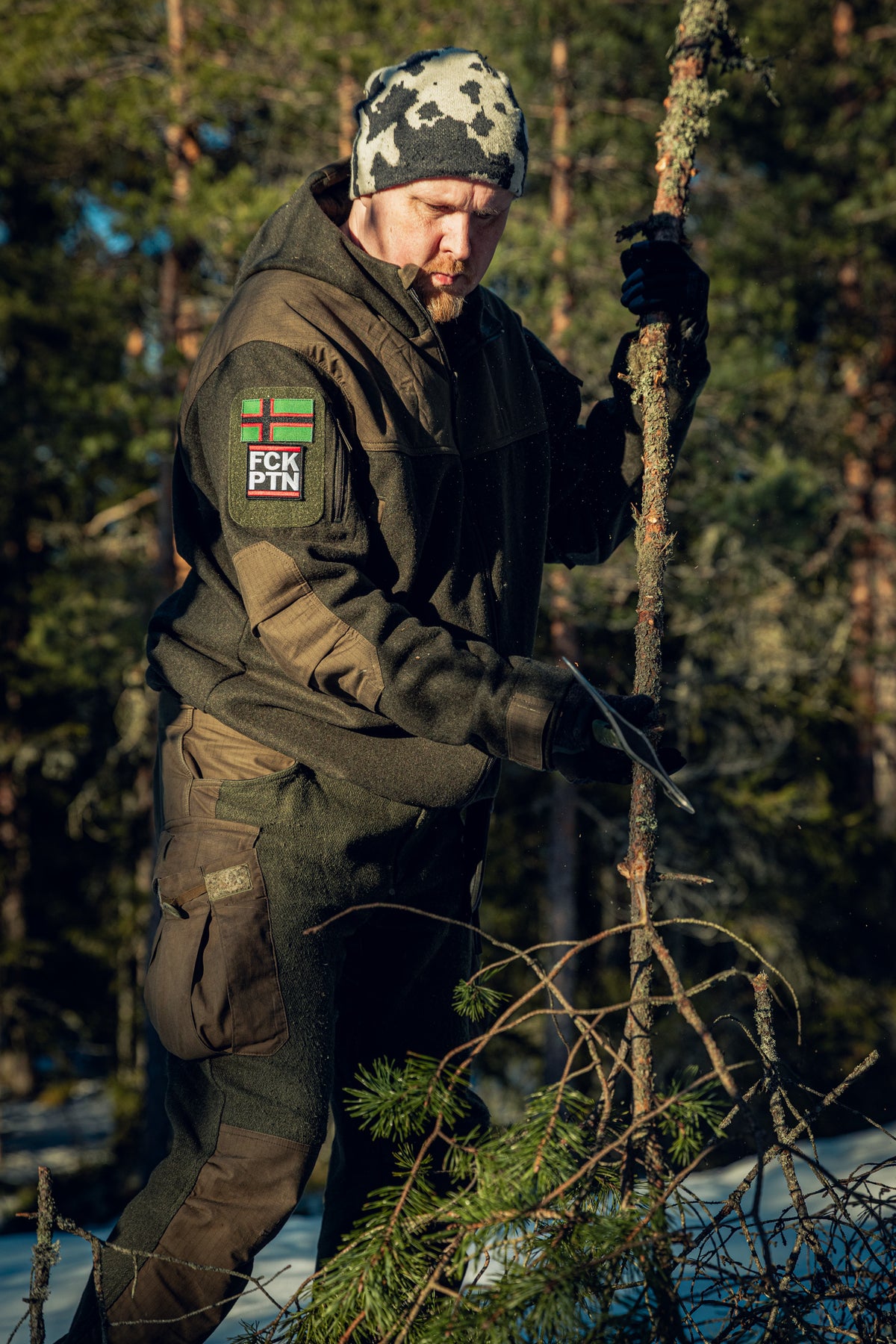 A man in a sunny pinewood forest with a pinewood branch in one hand and an axe in the other. There is still some snow on the ground, and he is wearing a green jacket and pants that appear to be made of wool, a black and white beanie, and black gloves.
