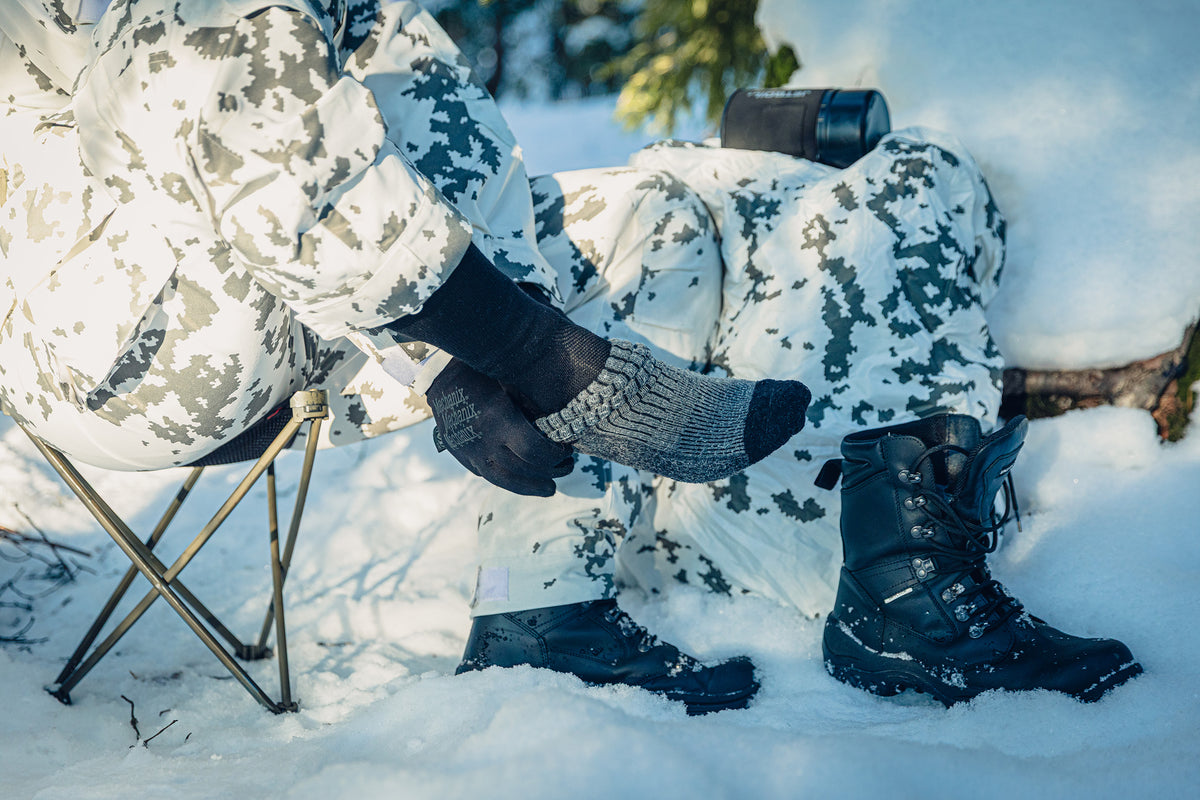 Eine Person sitzt auf einem Hocker im Schnee und zieht eine graue Socke über eine schwarze Socke. Die Person trägt Schneetarnkleidung.