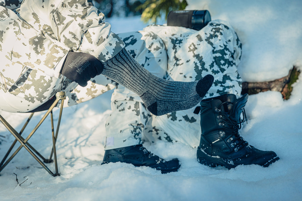 Nahaufnahme eines Fußes in einer grau gestreiften Socke. Die Person sitzt auf einem Hocker im Schnee, trägt Schneetarnkleidung und zieht mit behandschuhten Händen an der Socke.