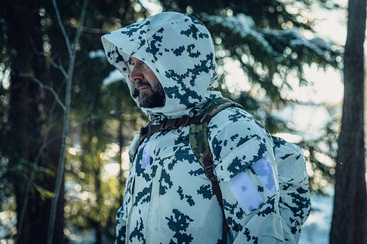 Ein Mann in einem sonnigen, verschneiten Wald trägt eine finnische M05-Schneetarnjacke mit aufgesetzter Kapuze und einen multicam-Rucksack. Er blickt nach links.