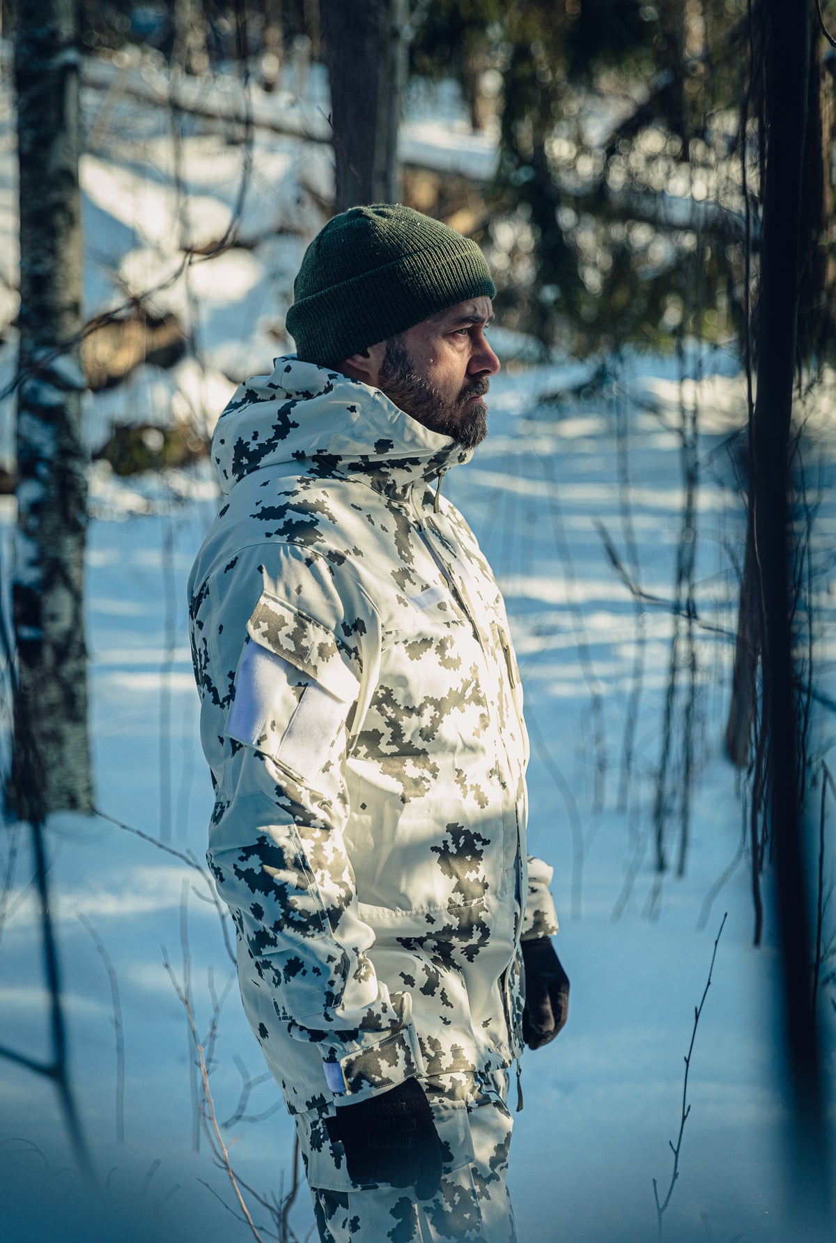 Ein Mann in einem sonnigen, verschneiten Wald trägt eine finnische M05-Schneetarnuniform mit Jacke und Hose sowie eine grüne Mütze. 