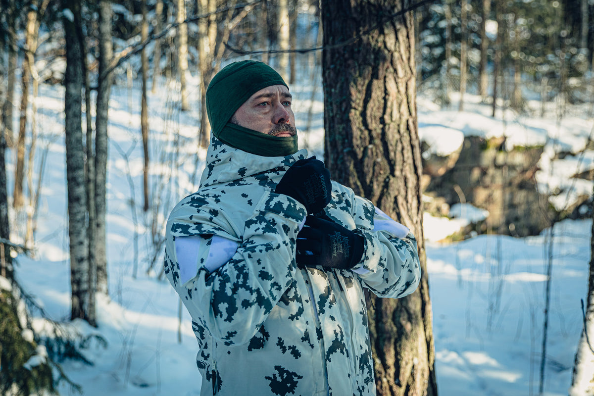 Ein Mann in einem sonnigen, verschneiten Wald trägt eine finnische M05-Schneetarnuniform und eine grüne Sturmhaube. Er stellt den Reißverschluss seiner Jacke ein.