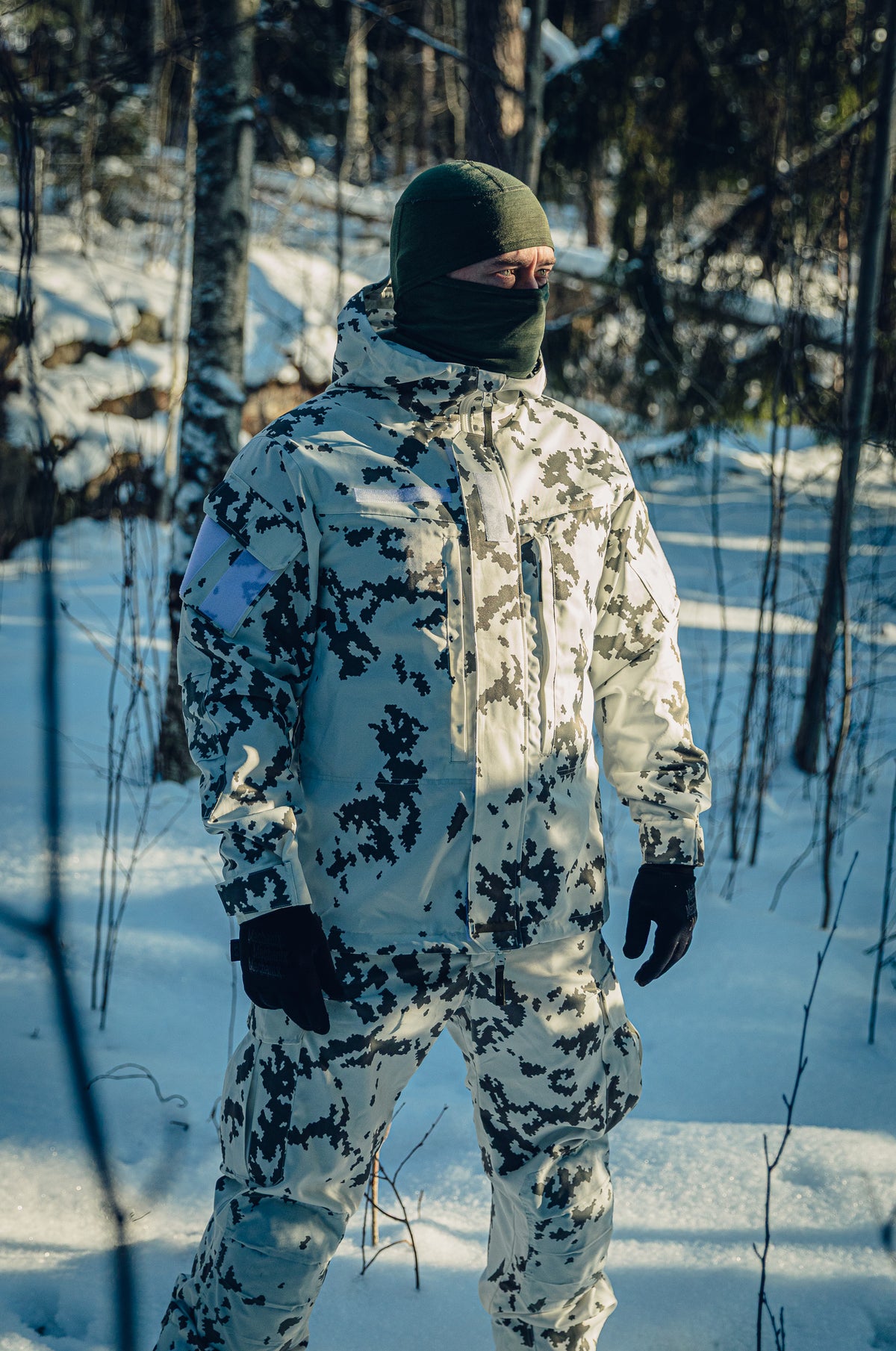 Ein Mann in einem sonnigen, verschneiten Wald trägt eine finnische M05-Schneetarnuniform und eine grüne Sturmhaube. Er blickt nach rechts in Richtung der Sonne.