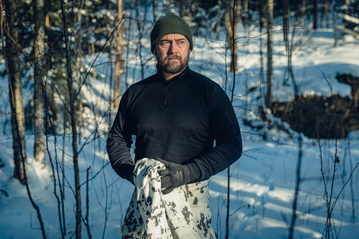 Ein Mann in einem sonnigen, verschneiten Wald trägt finnische M05-Schneetarnhose, ein schwarzes Rollkragenshirt und eine grüne Beanie. Er hält ein Schneetarn-Kleidungsstück in den Händen und schaut nach rechts.