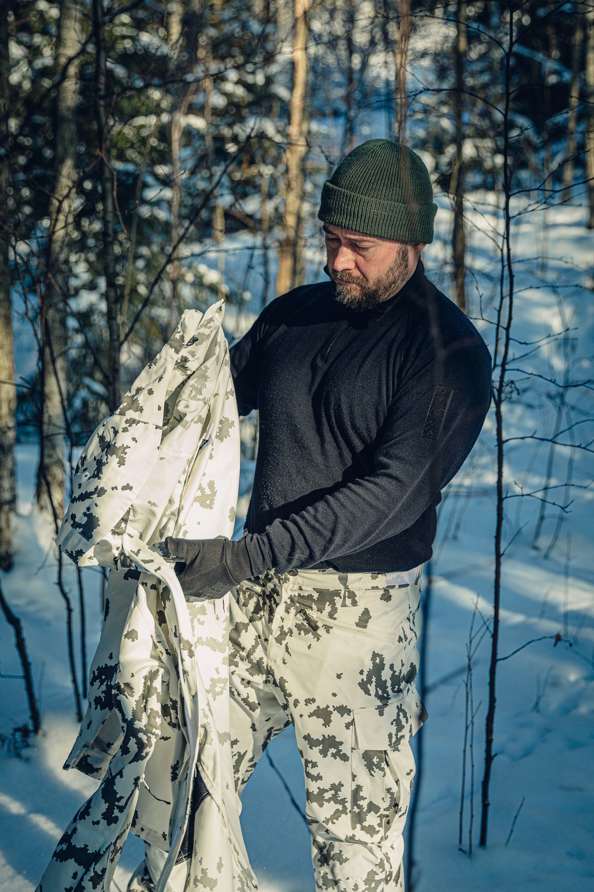 Ein Mann in einem sonnigen, verschneiten Wald trägt eine finnische M05-Schneetarnhose, ein schwarzes Poloshirt und eine grüne Beanie. Er hält eine Schneetarnjacke in den Händen und blickt darauf hinunter.
