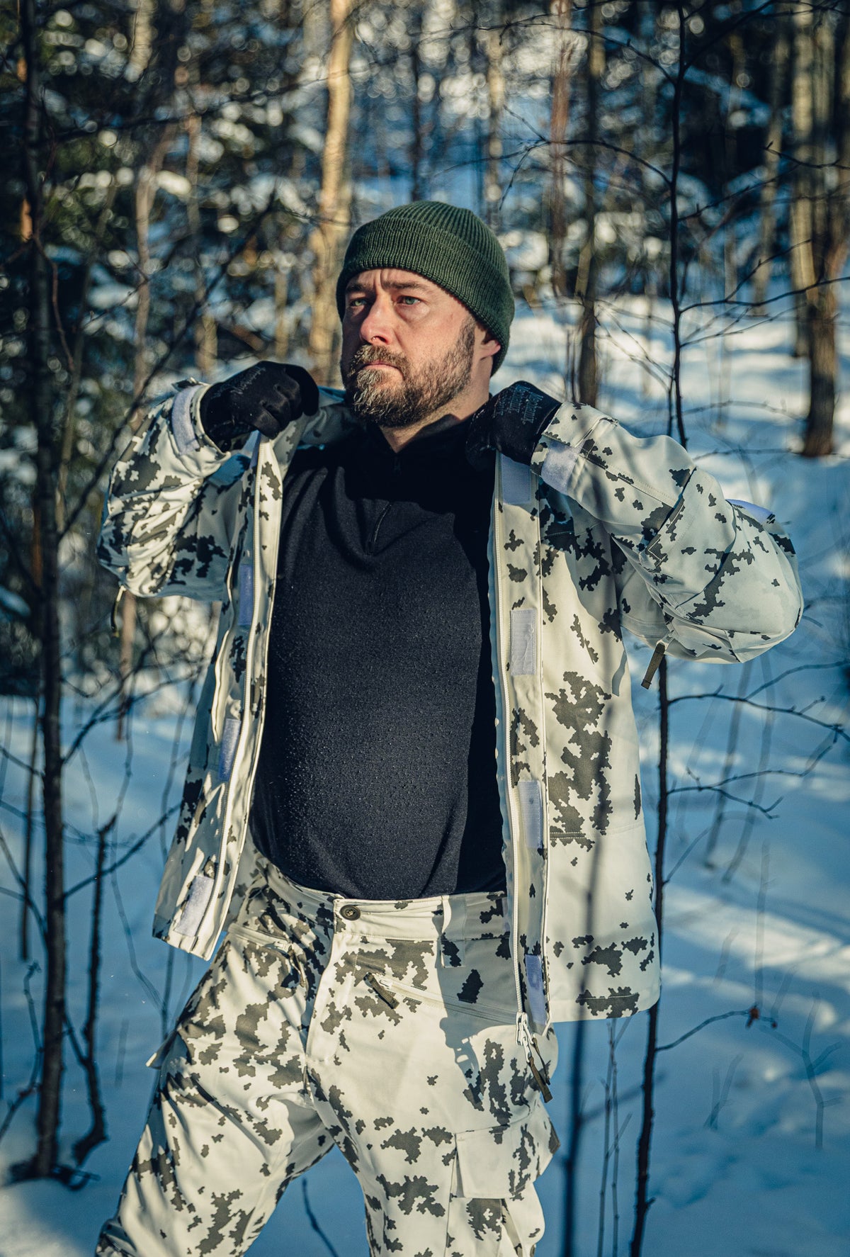 Ein Mann in einem sonnigen, verschneiten Wald trägt eine finnische M05-Schneetarnjacke und -hose, ein schwarzes Rollkragenshirt und eine grüne Beanie. Die Jacke ist offen, und er hält sie an den Seiten fest, als würde er sie zurechtrücken.
