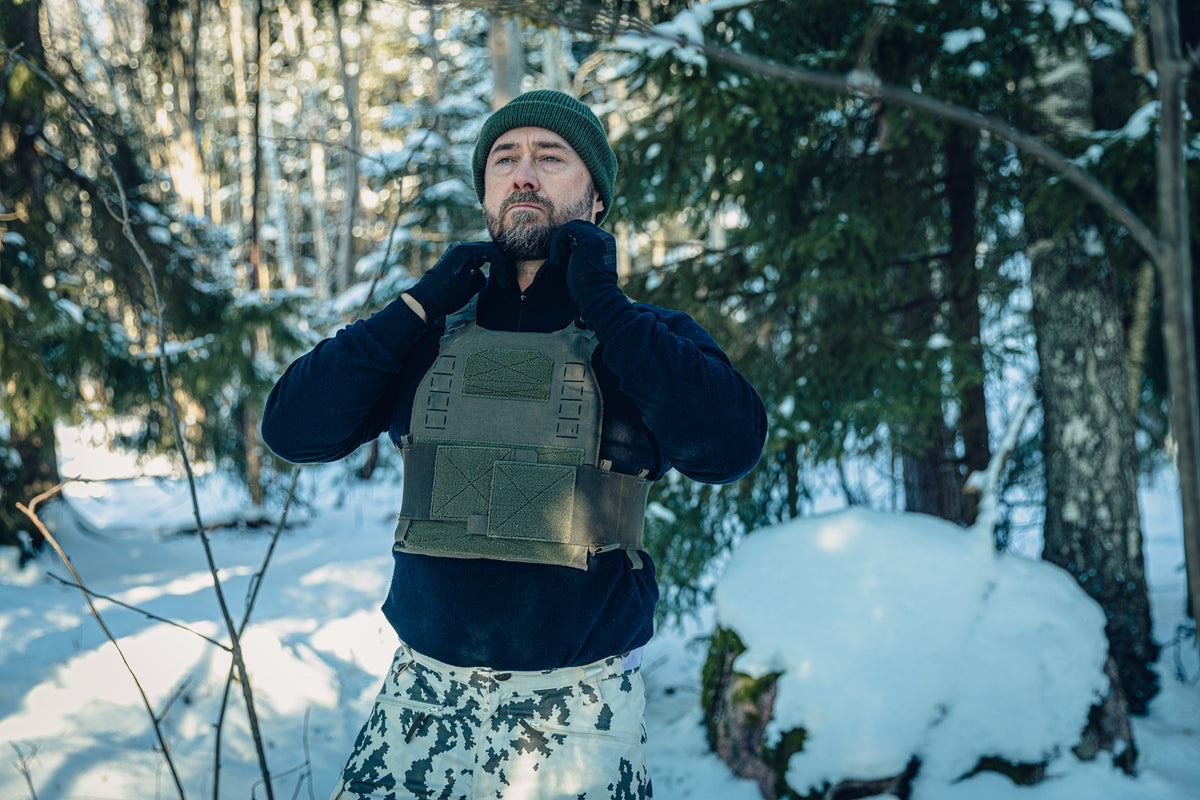 Ein Mann in einem sonnigen, verschneiten Wald trägt eine finnische M05-Schneetarnhose, ein schwarzes, langärmliges Rollkragenshirt, eine grüne Mütze und ein grünes plate carrier. Er richtet den Kragen seines Shirts.
