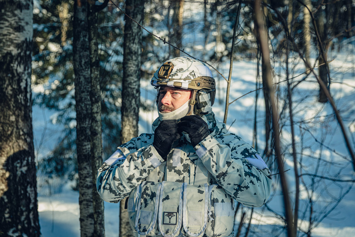 A man in a sunny, snowy forest wearing a Finnish M05 snow camo uniform and a combat helmet.  He is adjusting the strap of his helmet and there is a white neck tube covering part of his chin.