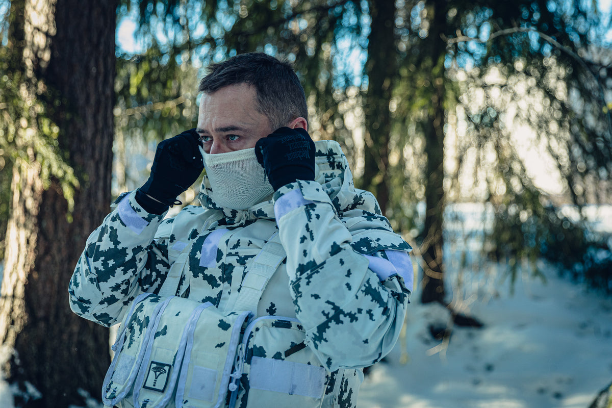 A man in a sunny, snowy forest wearing a Finnish M05 snow camo uniform. He is pulling a white neck tube to cover his nose.