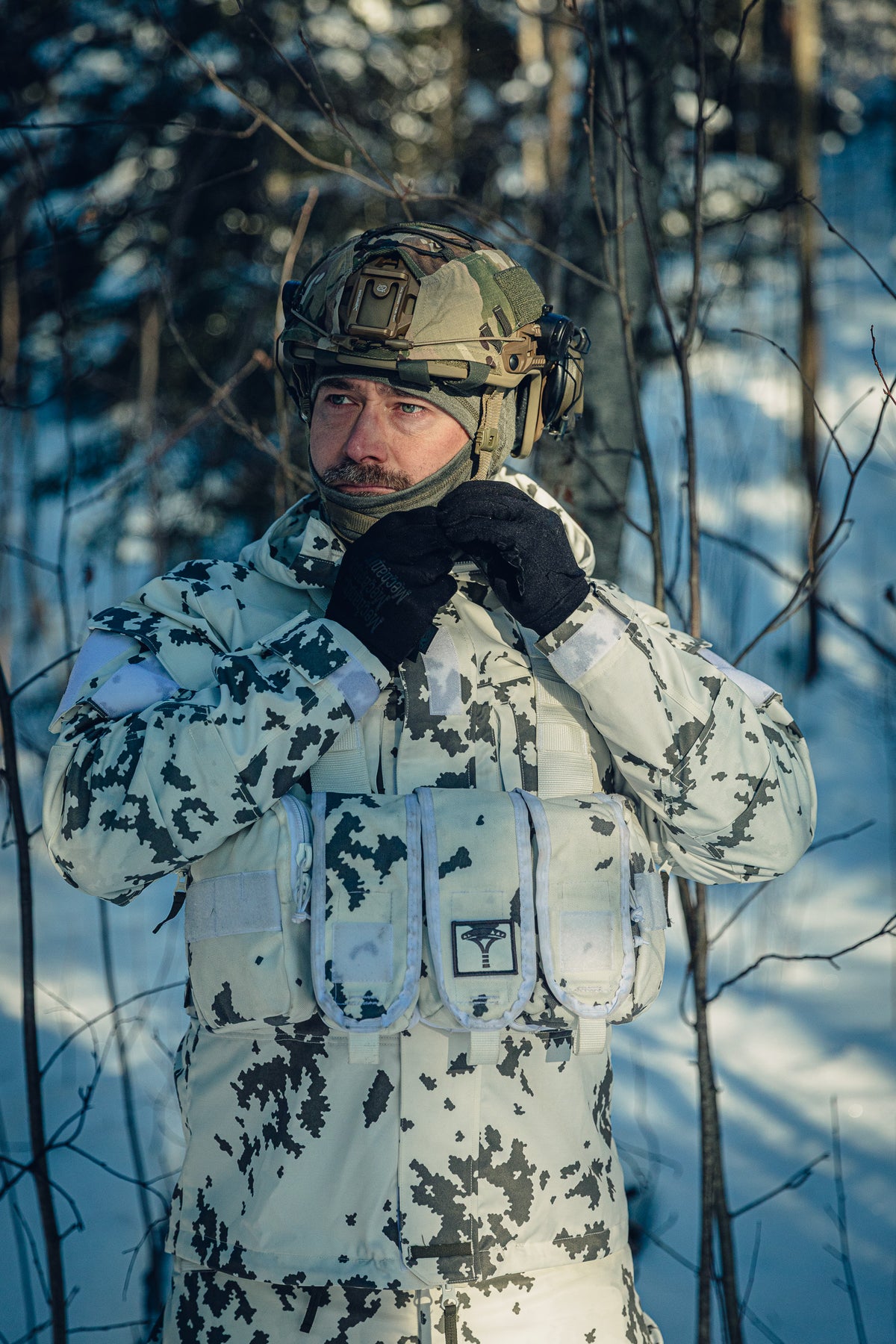 Ein Mann in einem sonnigen, verschneiten Wald trägt eine finnische M05-Schneetarnuniform und einen Gefechtshelm. Er stellt den Riemen seines Helms ein, und unter dem Helm trägt er eine graue Sturmhaube.