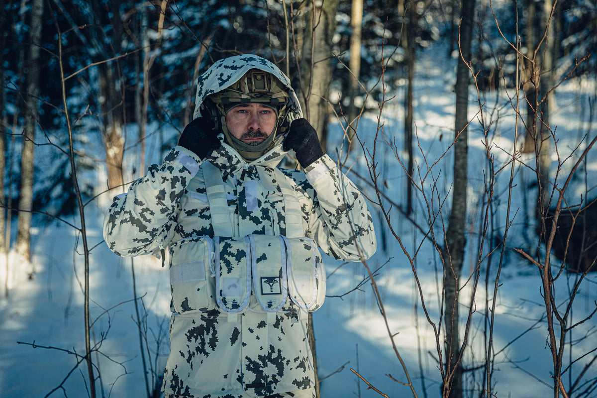 Ein Mann in einem sonnigen, verschneiten Wald trägt eine finnische M05-Schneetarnuniform und einen Gefechtshelm. Er richtet die Kapuze der Jacke, die über dem Helm liegt, und unter dem Helm trägt er eine graue Sturmhaube.
