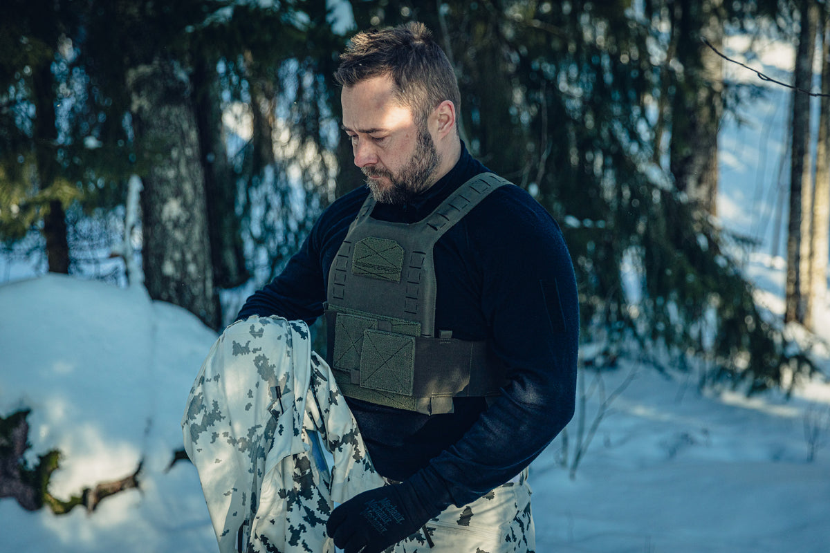 Ein Mann in einem sonnigen, verschneiten Wald trägt eine finnische M05-Schneetarnhose, ein schwarzes, langärmliges Poloshirt, ein grünes plate carrier. Er hält ein weiteres Schneetarn-Kleidungsstück in den Händen und schaut nach unten.