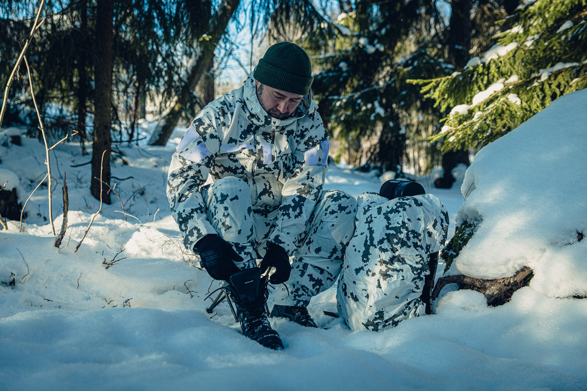 A man sitting on a stool in a snowy forest opening the laces of his combat boots. He is wearing Finnish M05 snow camo clothing, a green beanie, and there is a backpack next to him covered in a snow camo backpack cover.